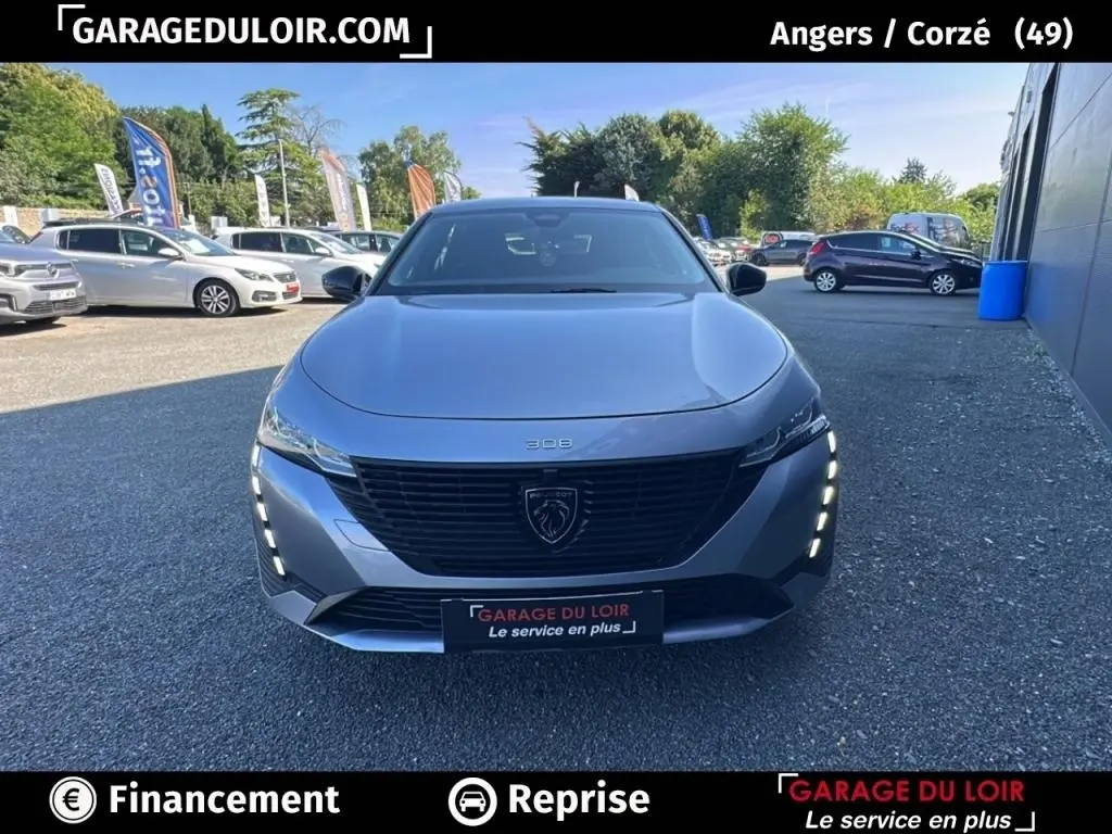 Vue avant frontale d'une Peugeot 308 gris clair 2023 avec feux LED verticaux allumés sur un parking extérieur.