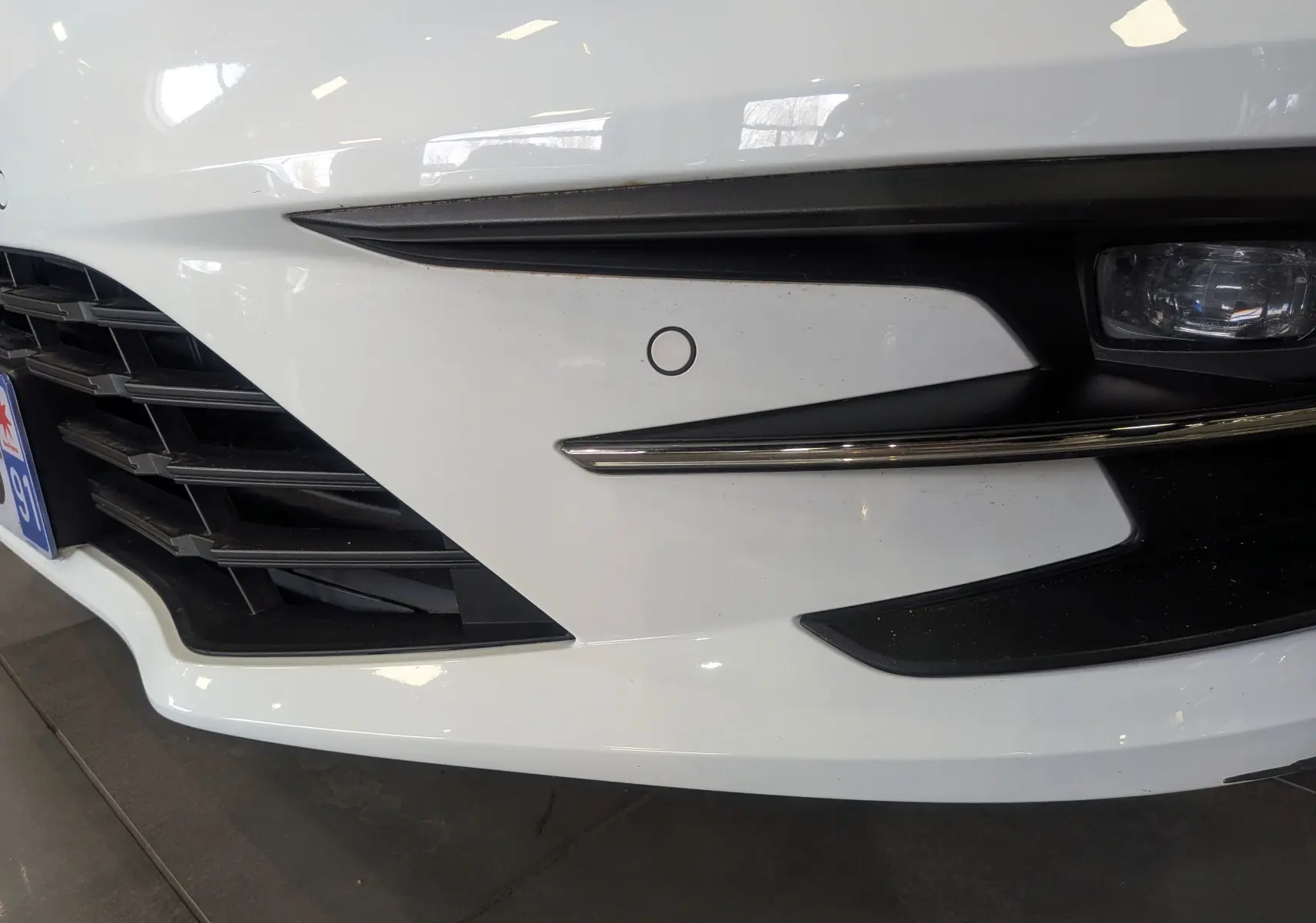 Gros plan sur le pare-chocs avant droit blanc glacier d’une Renault Mégane IV Berline avec radar de stationnement et antibrouillard.