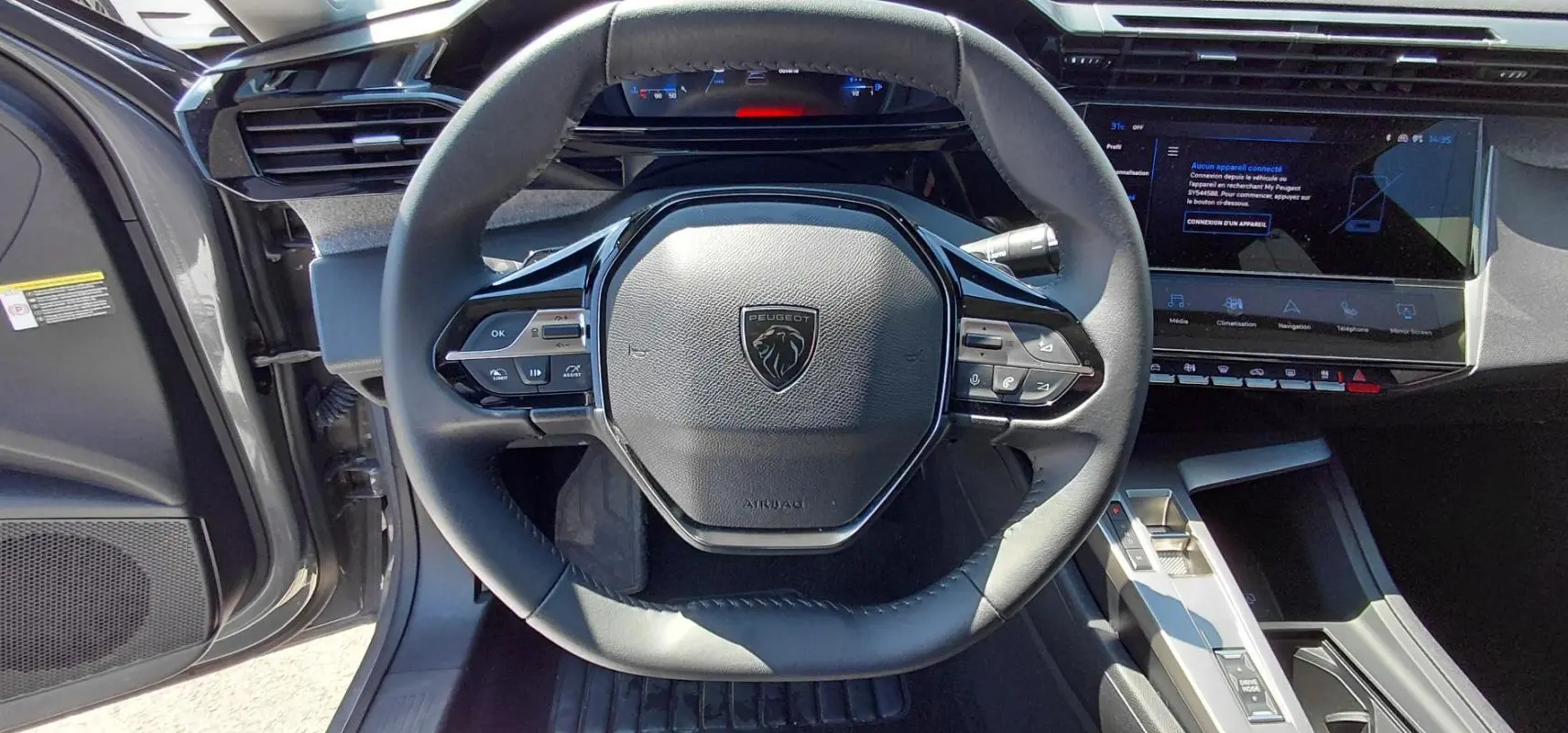 Vue intérieure centrée sur le volant cuir noir de la Peugeot 308 SW Hybrid 2025 avec tableau de bord numérique et écran tactile.