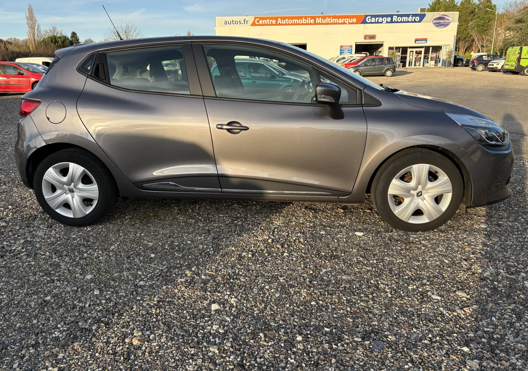 Profil côté gauche d'une Renault Clio IV gris métallisé, garée sur un sol gravillonné devant un garage multimarque.