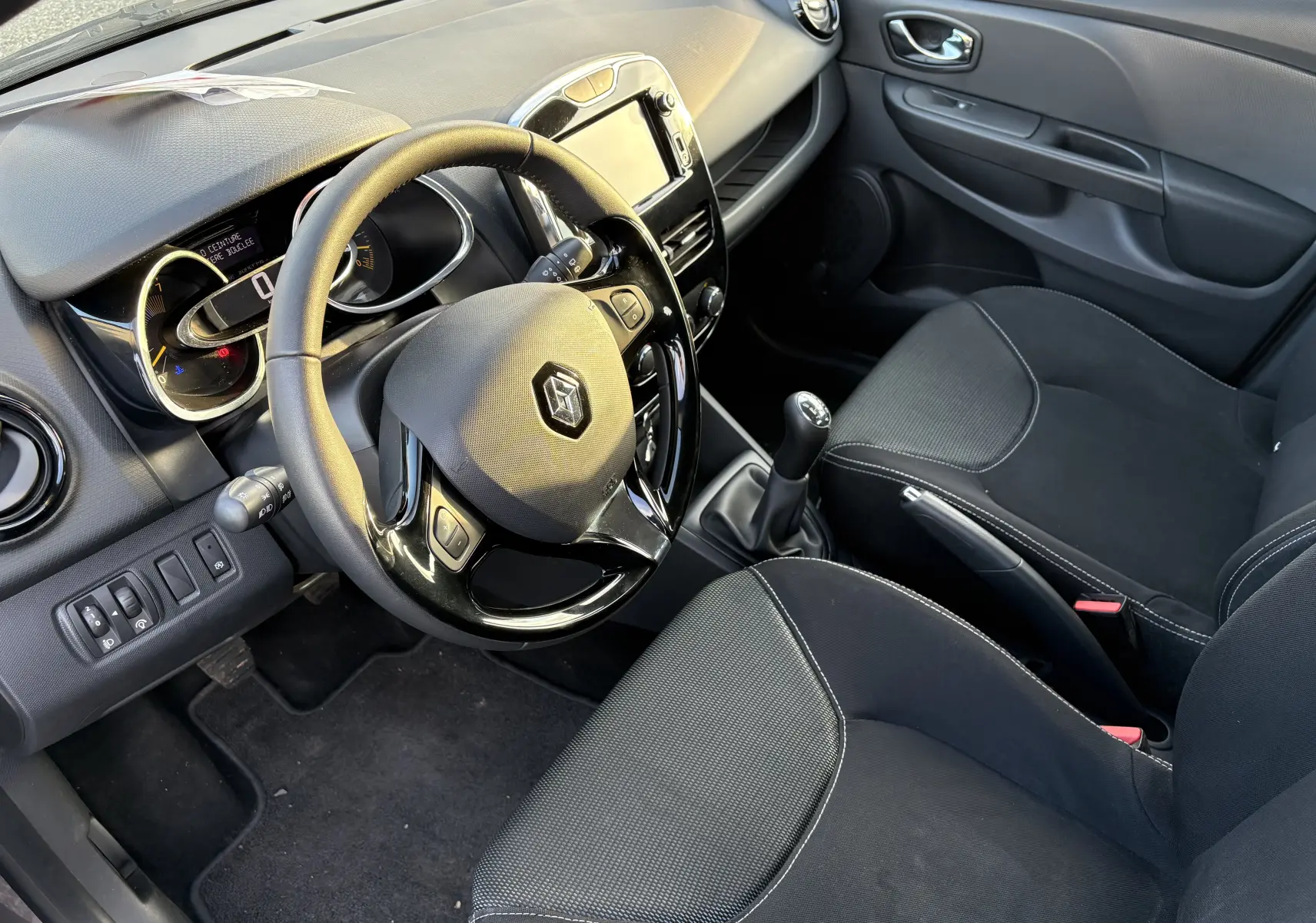 Vue intérieure côté conducteur d'une Renault Clio IV grise de 2013, avec volant, tableau de bord et GPS intégrés.