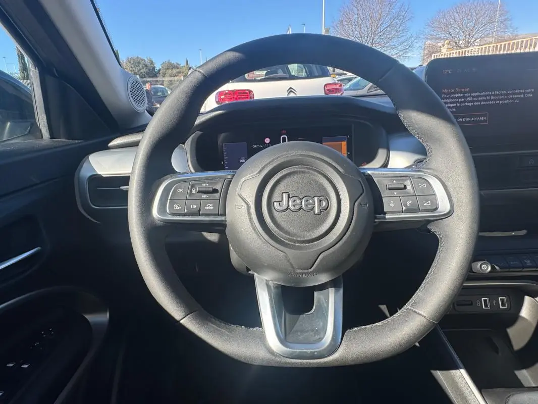 Vue intérieure centrée sur le volant Jeep noir avec commandes intégrées, tableau de bord et écran tactile visibles.