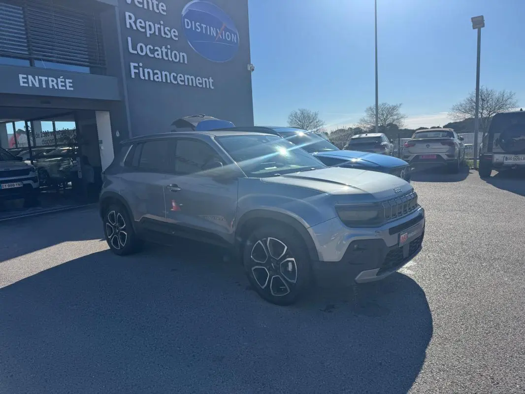 Jeep Avenger gris en 3/4 avant droit, stationné devant un bâtiment avec jantes alliage 18 pouces visibles.
