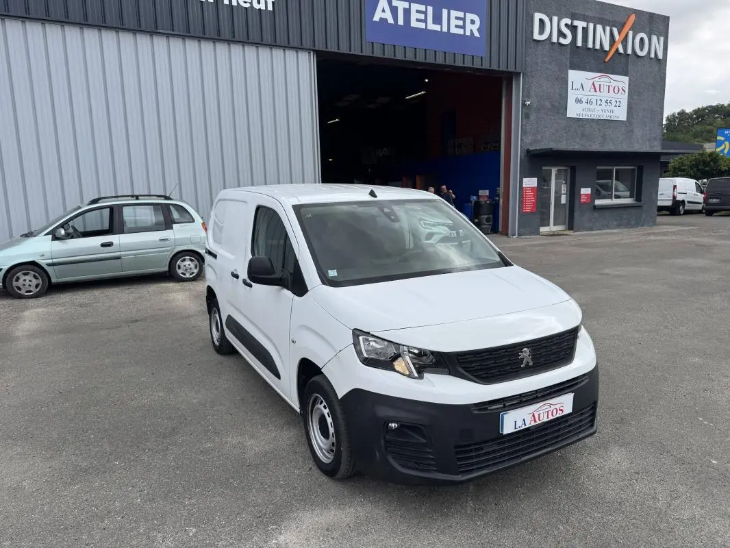 Peugeot Partner blanc vue 3/4 avant droit, avec pare-chocs noir, stationné devant un atelier automobile.