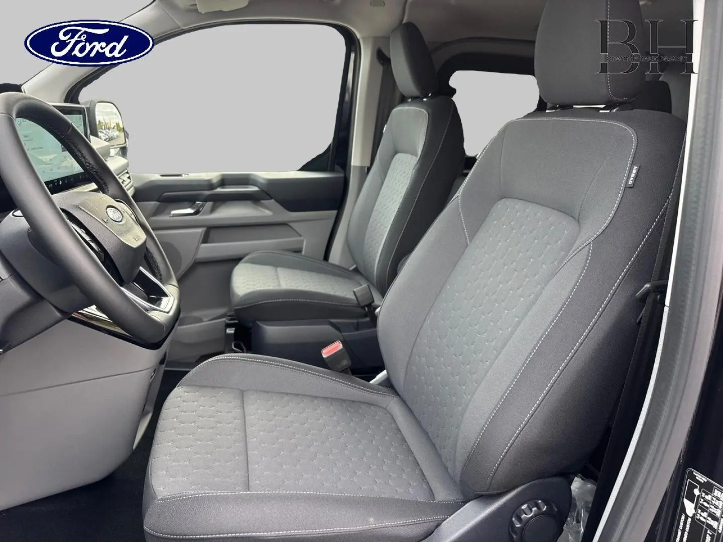 Vue intérieure côté conducteur du Ford Transit Custom Fourgon 2026, sièges tissu gris avec volant multifonction noir.