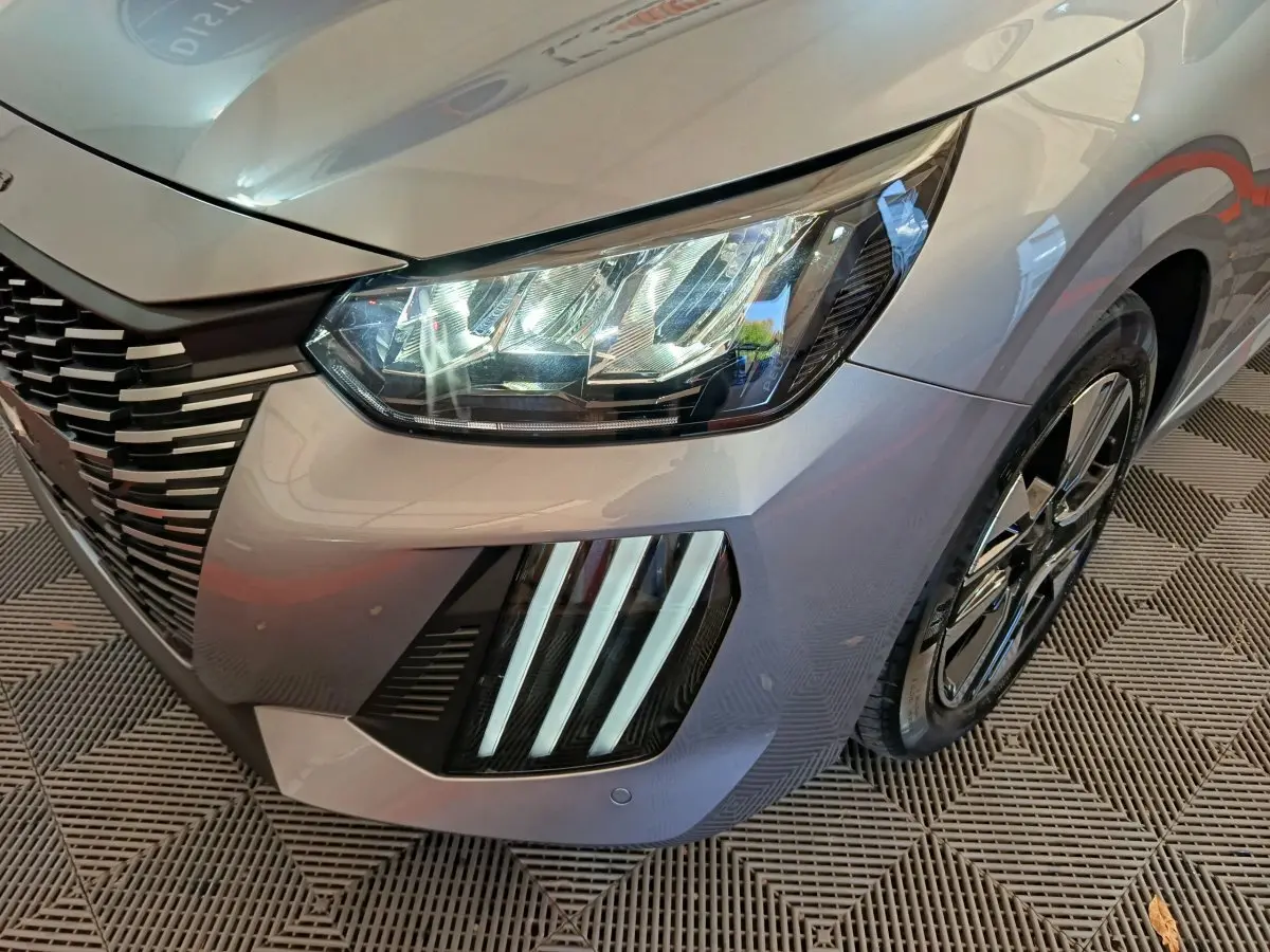 Gros plan sur l'avant droit gris clair d'une Peugeot 208 2025, mettant en valeur ses phares LED et la calandre distinctive.