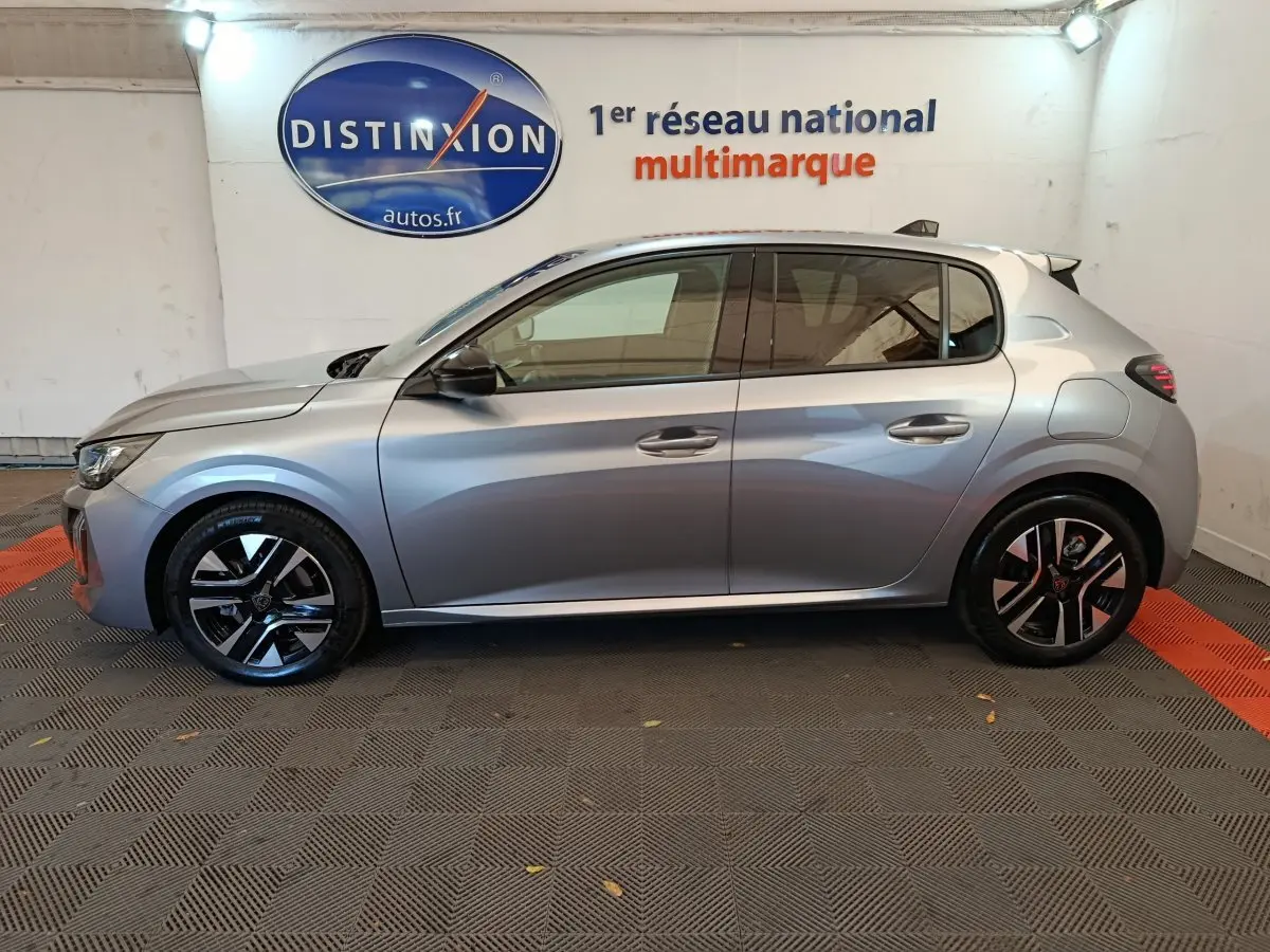 Profil côté gauche d'une Peugeot 208 PureTech 100 S&S Allure gris clair, en intérieur sur sol carrelé.