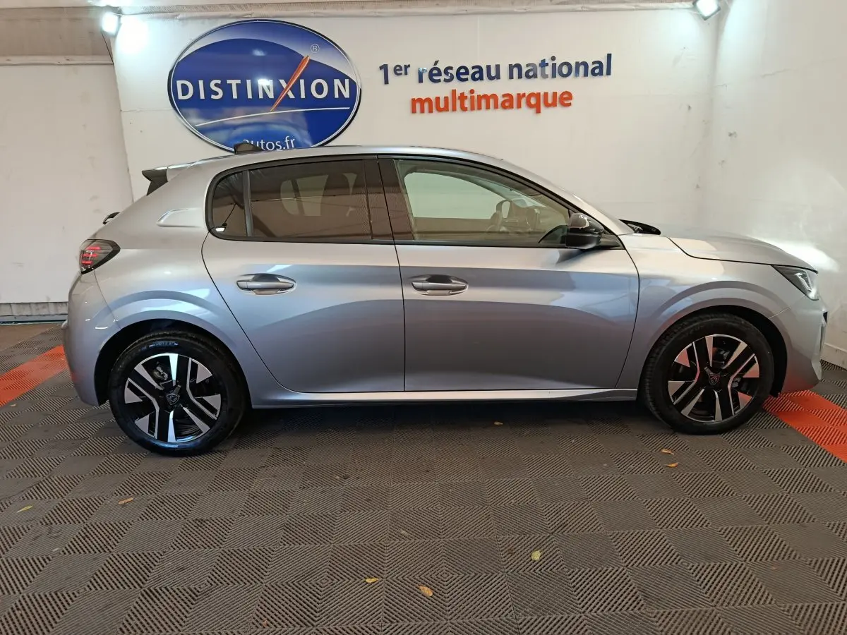 Profil côté droit d'une Peugeot 208 gris clair 2025 dans un showroom avec jantes noires et logo Distinxion en arrière-plan.