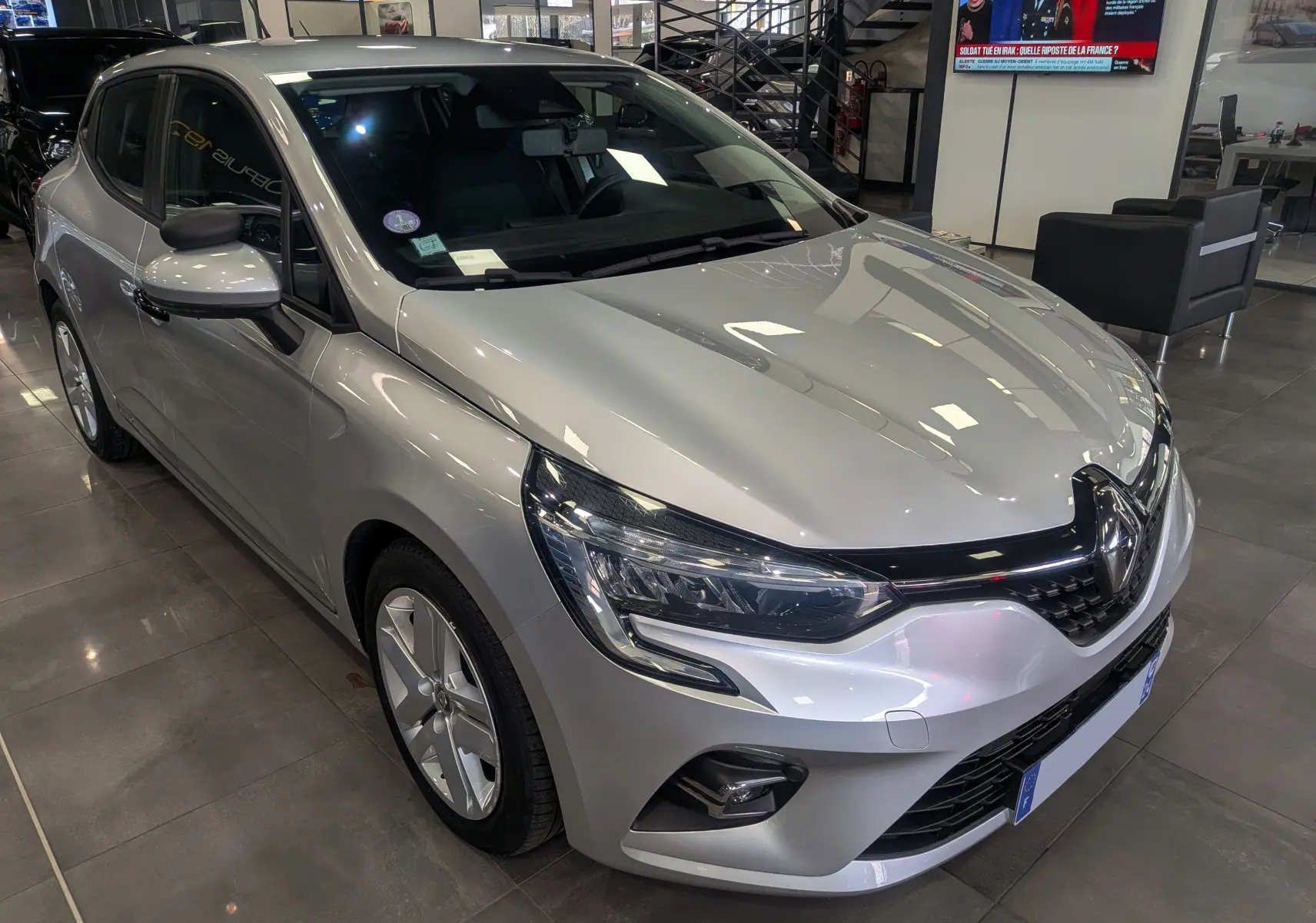 Renault Clio V gris platine vue 3/4 avant droit en showroom, avec phares LED et calandre noire brillante.