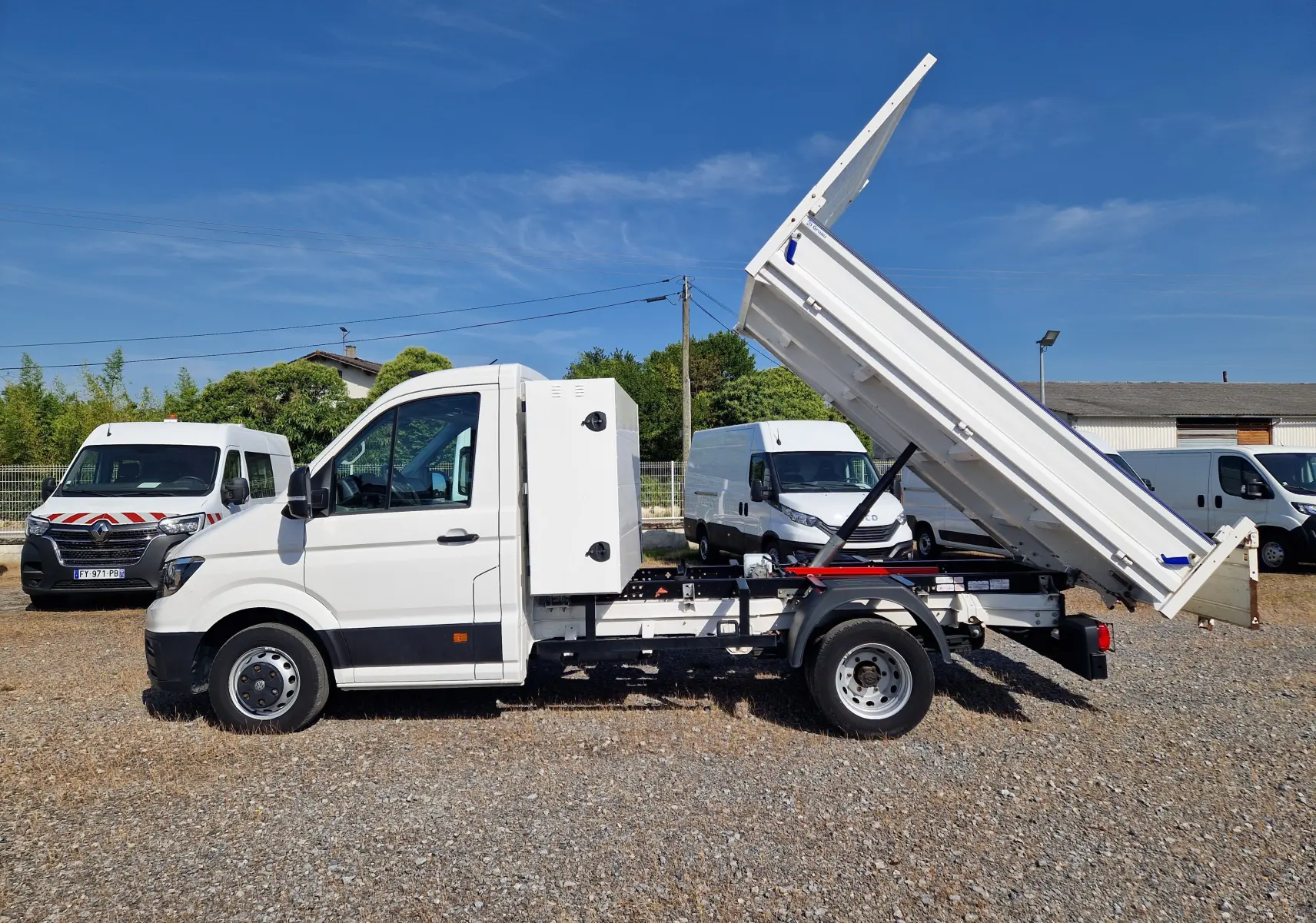 Vue de profil droit d’un Volkswagen Crafter blanc avec benne relevée et coffre latéral sur un terrain extérieur.