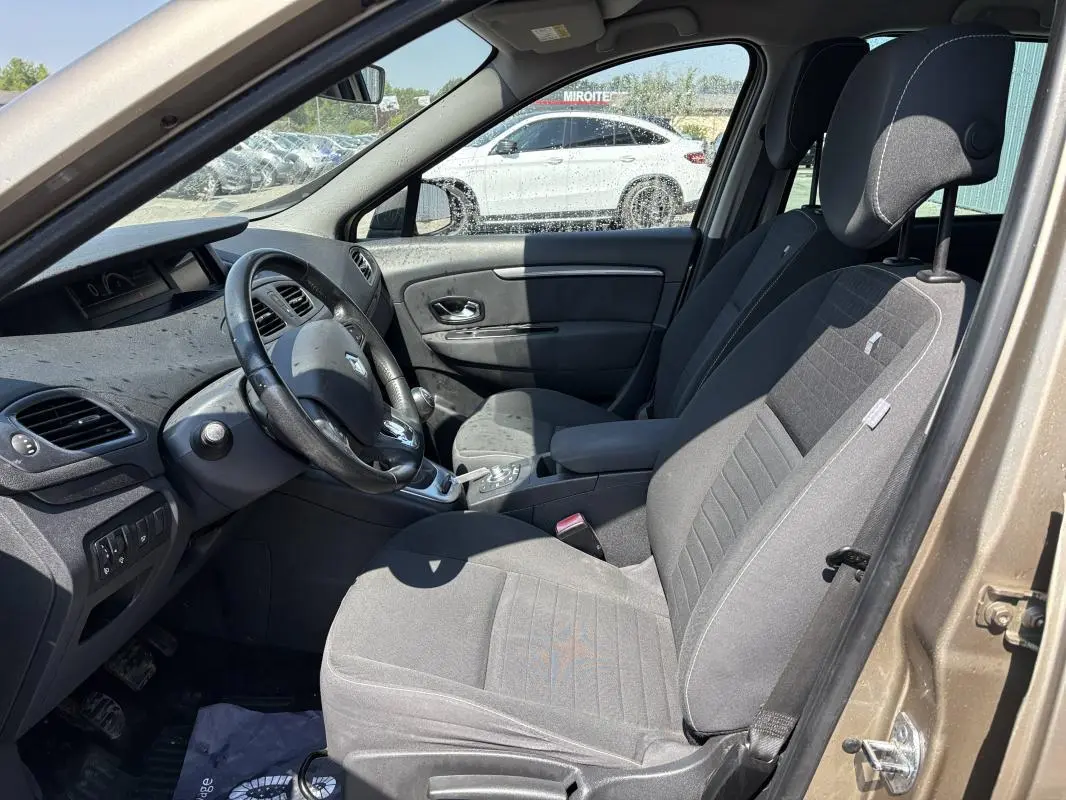 Vue intérieure côté conducteur d’un Renault Scénic III beige 2015, avec volant cuir et accoudoir central visible.
