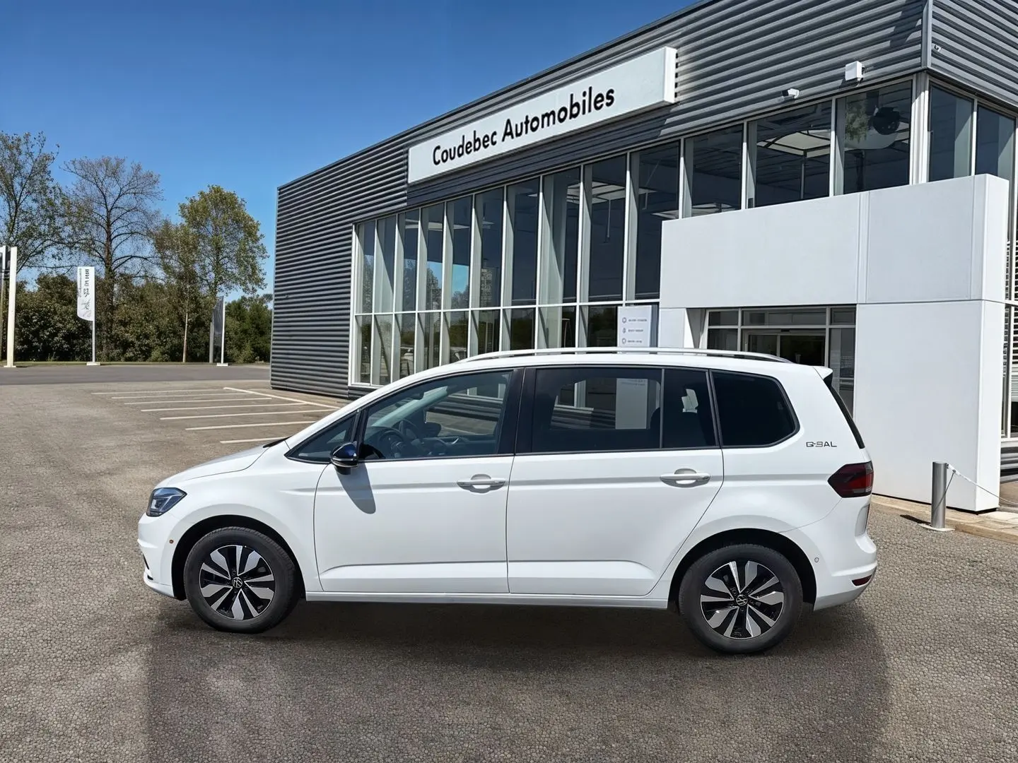 Vue de profil côté gauche d’un Volkswagen Touran blanc 2025 garé devant un bâtiment moderne de concession.