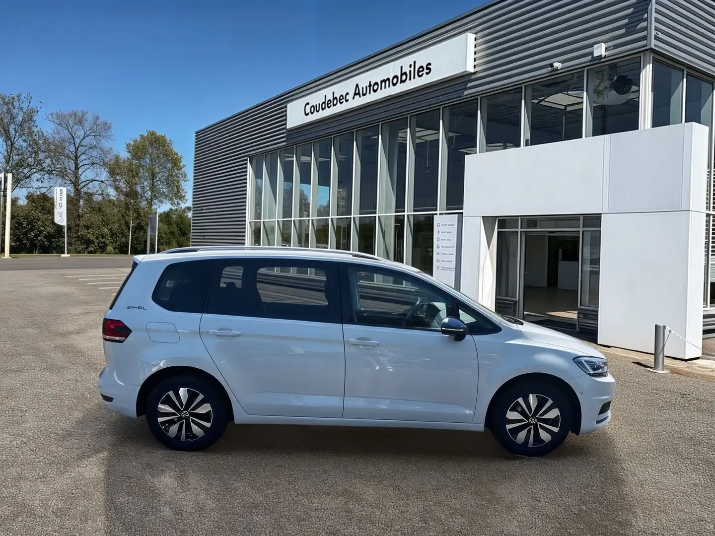 Profil côté gauche d'un Volkswagen Touran blanc 2025 stationné devant un bâtiment moderne de concession.