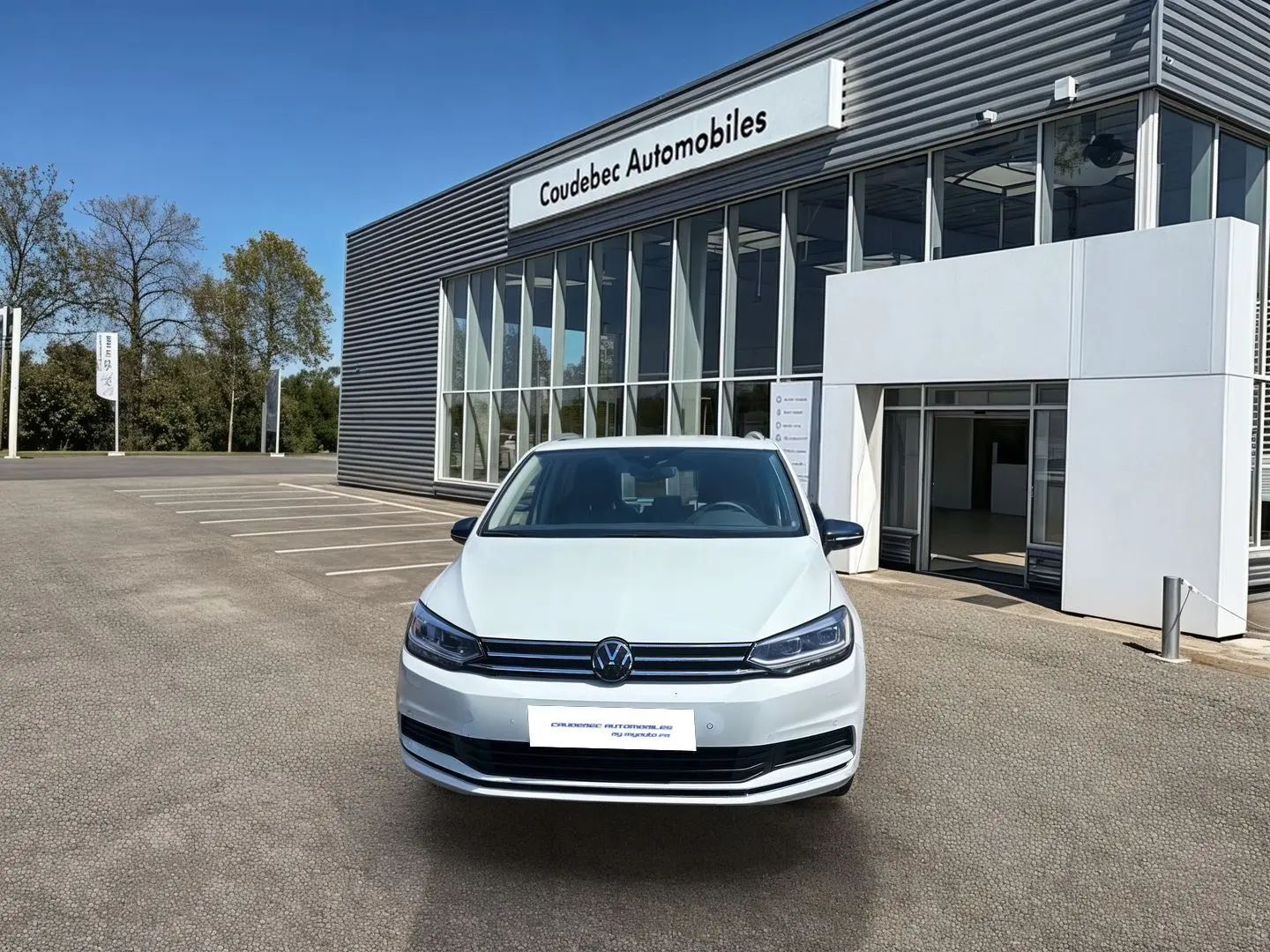 Volkswagen Touran blanc vue de face, stationné devant un bâtiment commercial sous un ciel bleu.