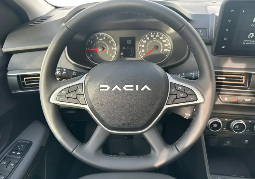 Vue rapprochée du volant noir Dacia avec commandes intégrées, tableau de bord analogique et écran tactile central.