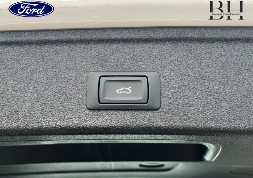 Gros plan sur le bouton d'ouverture électrique du coffre, entouré de garniture noire texturée.