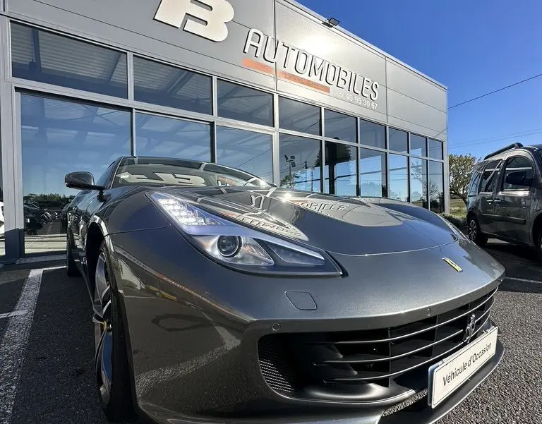 Ferrari GTC4 Lusso gris foncé vue 3/4 avant droit, avec phares allumés devant un showroom FB Automobiles.