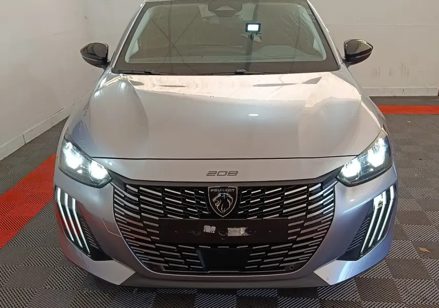 Vue frontale d'une Peugeot 208 gris clair 2025 avec phares LED et calandre imposante en intérieur showroom.