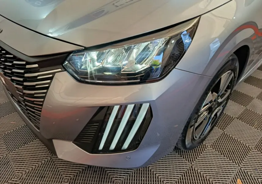 Gros plan sur l'avant droit gris clair de la Peugeot 208 2025 avec phares LED et signature lumineuse verticale distinctive.