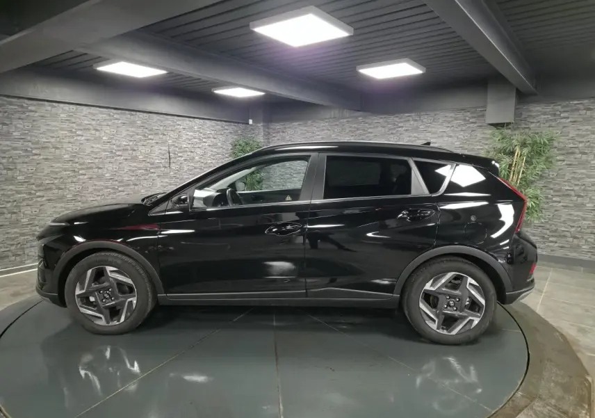 Vue de profil côté gauche d'une Hyundai Bayon noire métallisée 2025 dans un showroom avec jantes alliage 17 pouces.