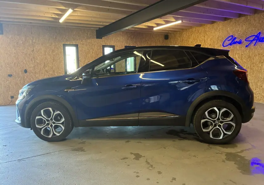Vue de profil côté gauche d'un Renault Captur bleu 2021 avec toit noir et jantes alliage distinctives dans un intérieur en bois.