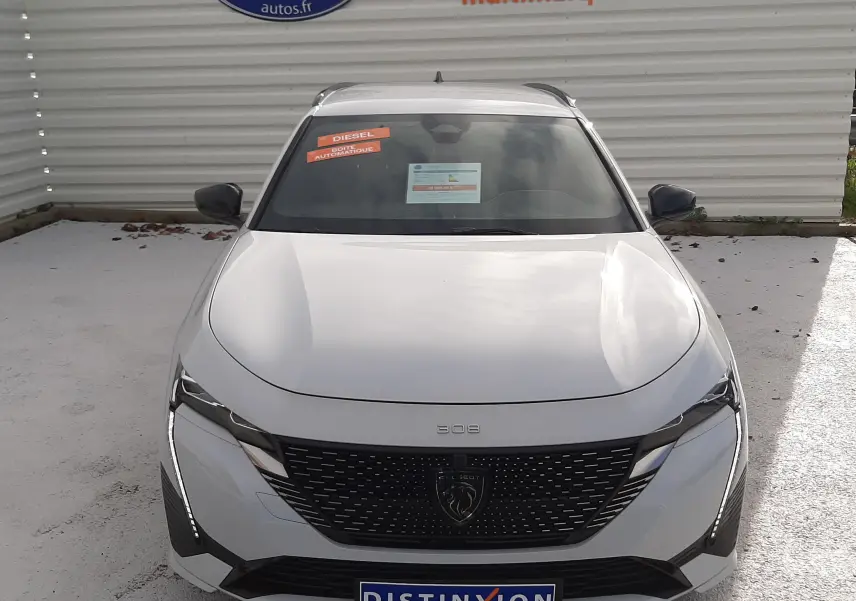 Vue avant d'une Peugeot 308 SW blanche Okenite avec calandre noire et phares LED, stationnée devant un mur publicitaire.