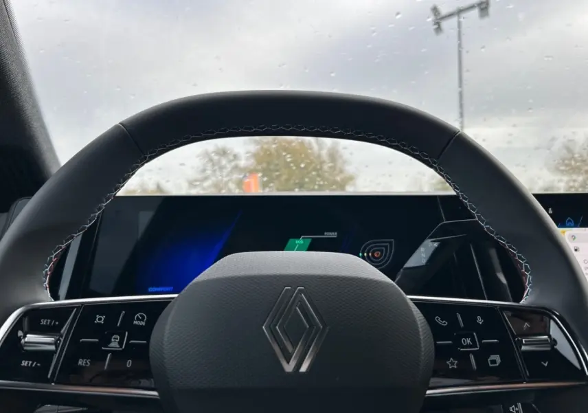 Volant noir avec logo Renault en gros plan, tableau de bord digital lumineux visible derrière sous pluie légère.