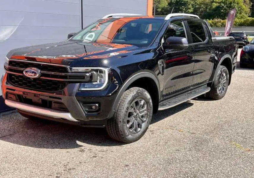 Ford Ranger Wildtrak Plus noir agate vu en 3/4 avant droit, avec jantes alu et barres de toit visibles.