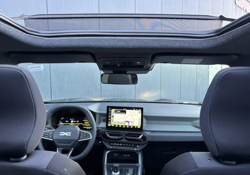 Vue intérieure avant du Dacia Bigster 2026 noir nacré, avec tableau de bord numérique et toit panoramique ouvert.