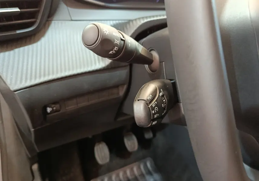 Gros plan sur les commandes au volant côté gauche de la Peugeot 208 gris clair, avec levier d'éclairage et régulateur de vitesse.