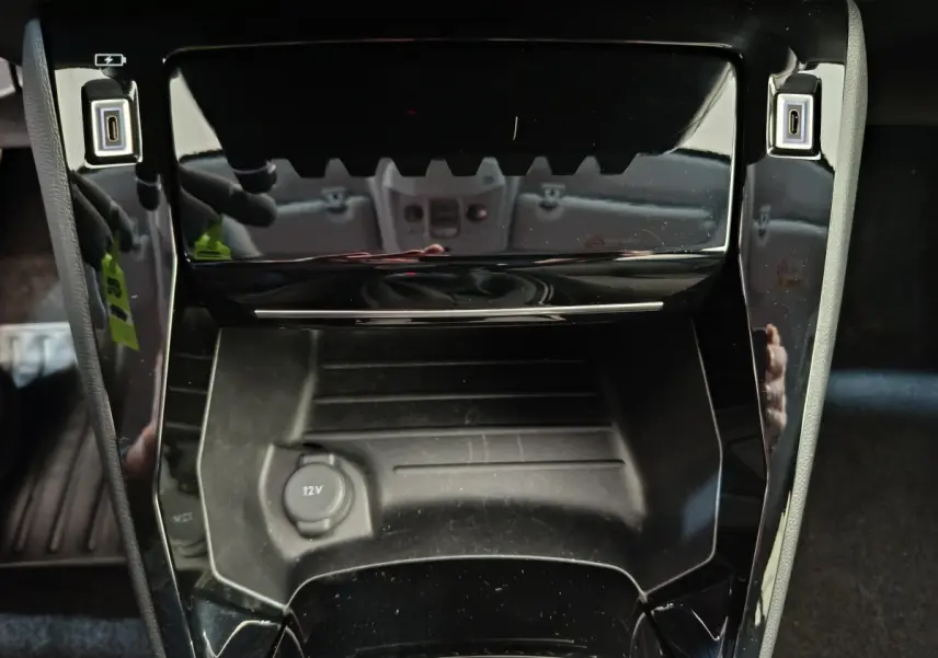 Gros plan sur la console centrale noire brillante de la Peugeot 208 PureTech 100 S&S Allure 2025 avec prise 12V.