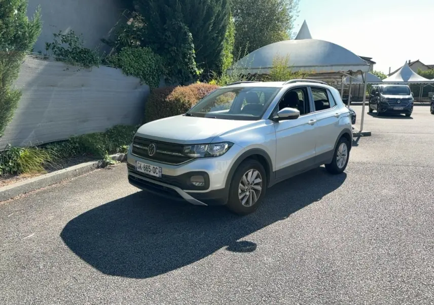 Volkswagen T-Cross gris clair en 3/4 avant droit, stationné sur un parking ensoleillé avec végétation en arrière-plan.