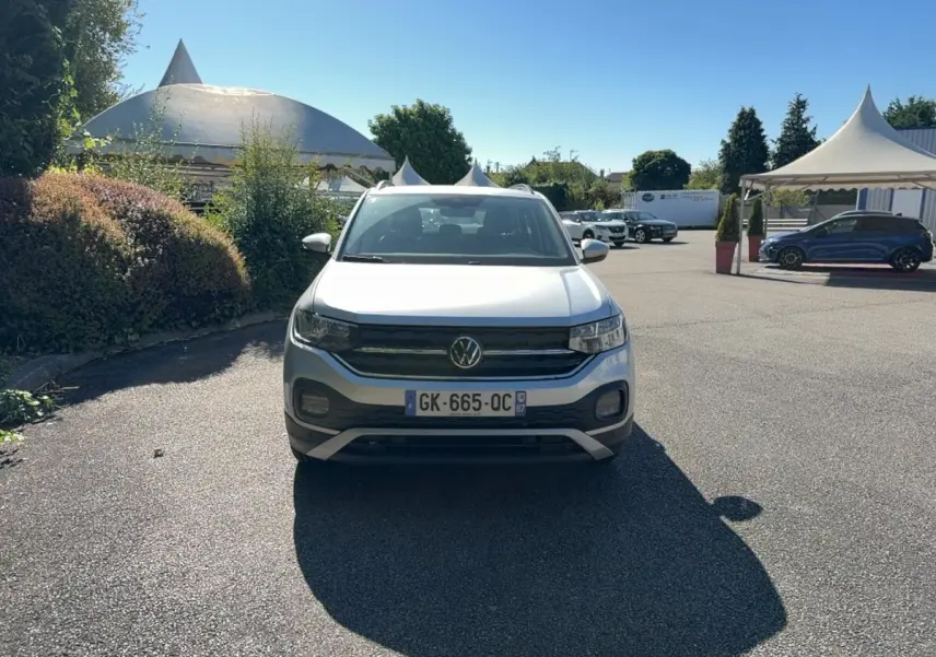 Volkswagen T-Cross gris clair vue de face, avec calandre noire et feux LED, stationné en extérieur sous un ciel bleu.