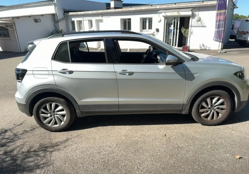 Vue de profil côté gauche d’un Volkswagen T-Cross gris clair avec jantes alliage et barres de toit noires.