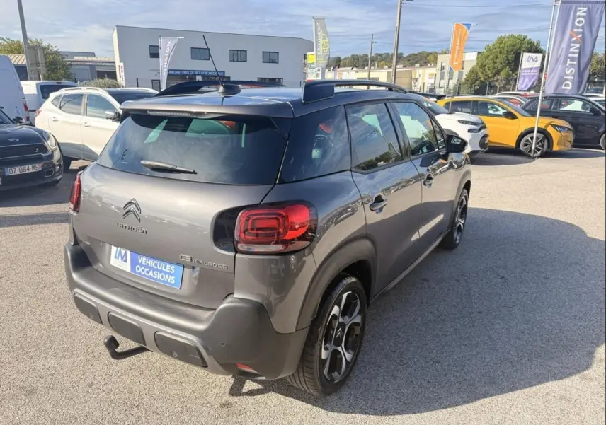 Vue 3/4 arrière droite d'un Citroën C3 Aircross gris métal 2021 avec barres de toit et attelage visible sur parking.