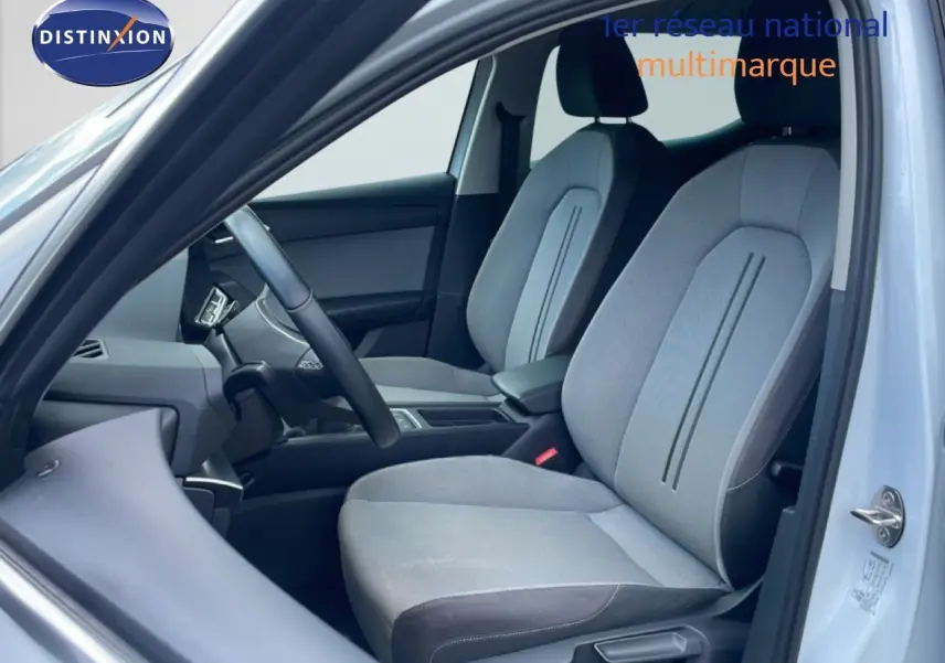 Vue intérieure côté conducteur d'une SEAT LEON 2025 blanc neige métal, sièges tissu gris clair avec détails noirs.