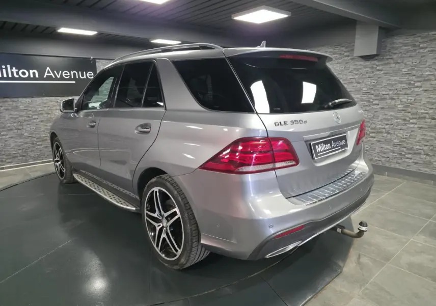 Vue 3/4 arrière droite d'un Mercedes GLE 350 d gris clair avec jantes AMG et attelage démontable en intérieur showroom.