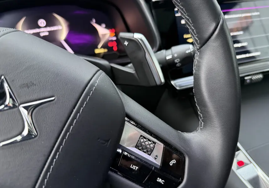 Gros plan sur le volant en cuir noir du DS7 2025, avec commandes intégrées et tableau de bord numérique flou en arrière-plan.