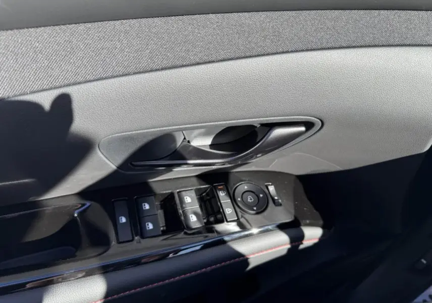 Gros plan sur la commande de lève-vitres et poignée intérieure côté conducteur du Hyundai Tucson noir avec surpiqûres rouges.