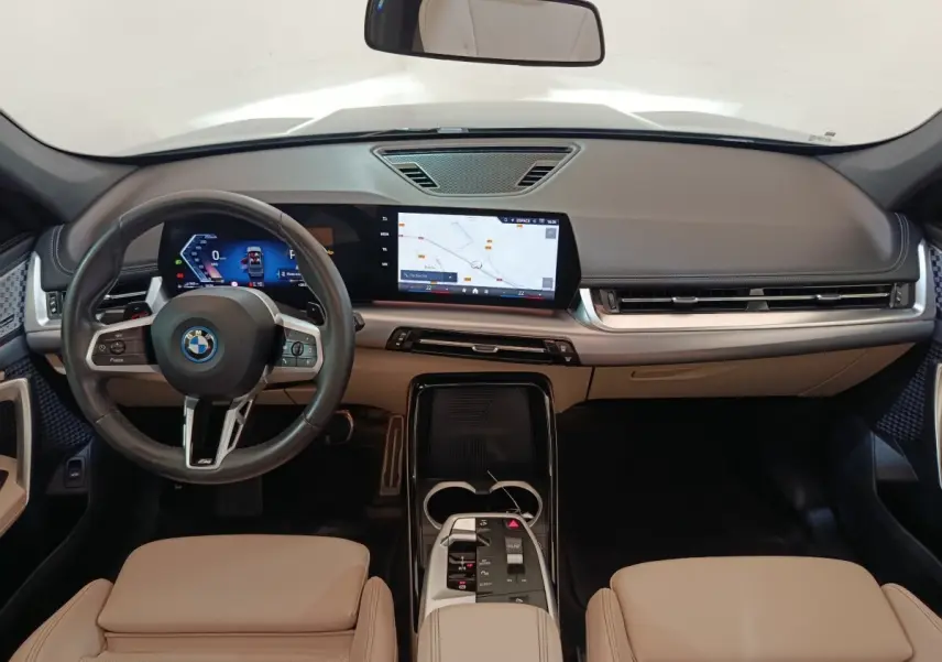 Vue intérieure avant du BMW X1 xDrive25e M Sport 2024 avec tableau de bord beige et écran tactile central lumineux.