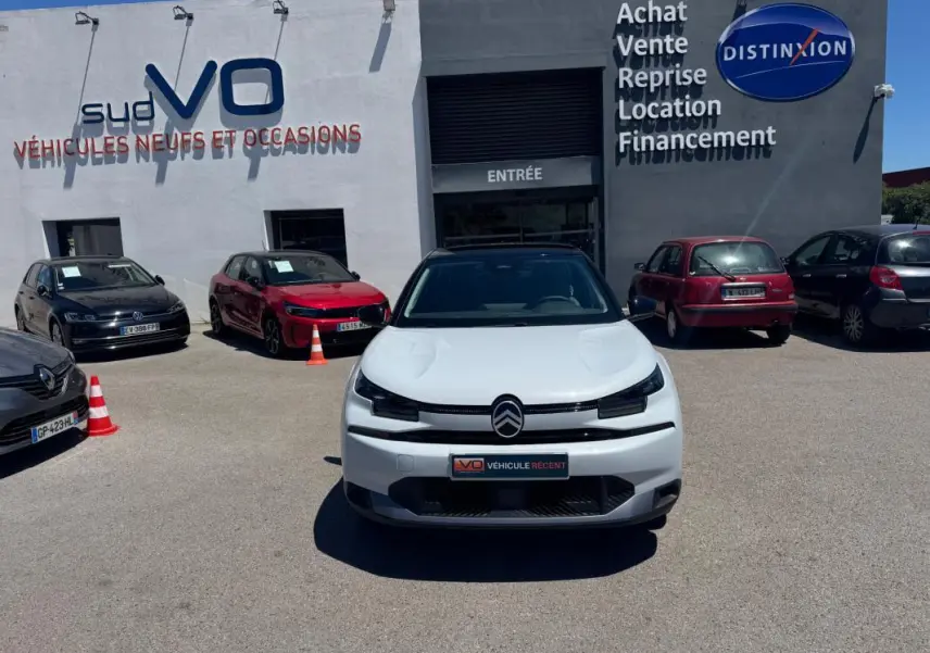 Citroën C4 hybride blanche vue de face, avec calandre noire et logo centré, stationnée devant un garage.