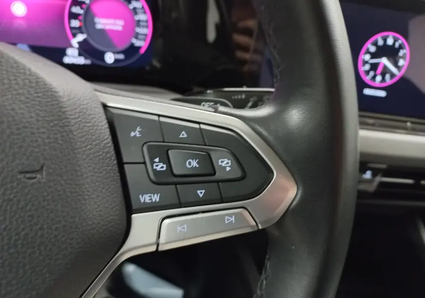 Gros plan sur les commandes au volant côté gauche d’une Volkswagen Golf 2023, avec tableau de bord numérique violet.