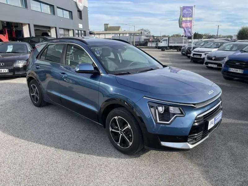 KIA Niro 2024 bleu métal en 3/4 avant droit, avec signature lumineuse LED et jantes alliage distinctives.