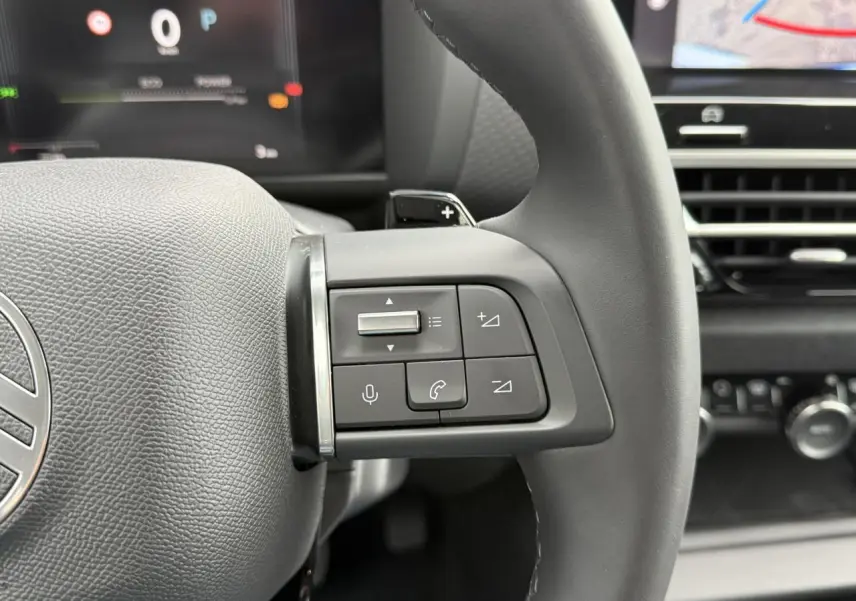 Gros plan sur les commandes au volant noir mat du Citroën C4 hybride 2025 avec écran numérique en arrière-plan.