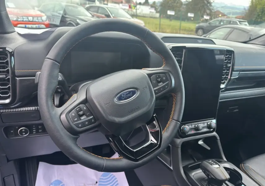 Vue intérieure centrée sur le volant cuir noir avec surpiqûres orange du Ford Ranger 2025, écran tactile vertical et console centrale.