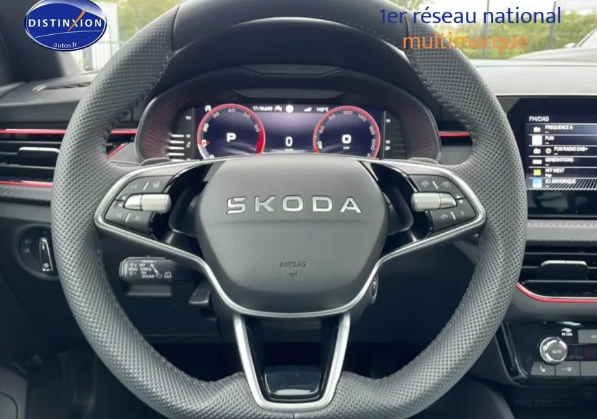 Vue centrée sur le volant perforé SKODA avec tableau de bord numérique et touches multifonctions, ambiance intérieure noire avec touches rouges.