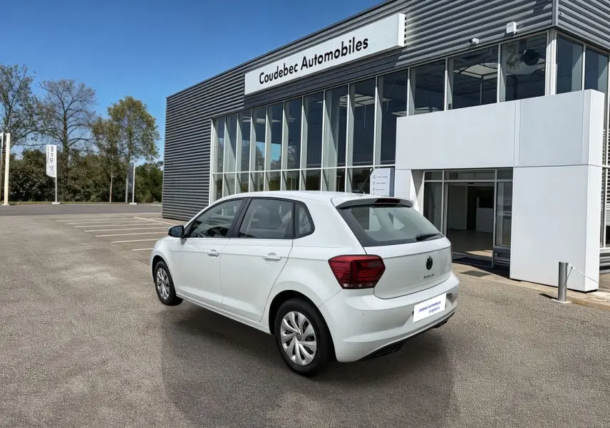 Volkswagen Polo Business blanche vue 3/4 arrière droit, avec jantes acier et feux arrière rouges, devant un garage.