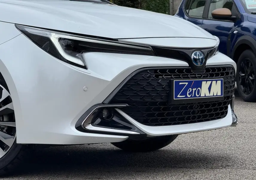 Gros plan sur l'avant droit blanc lunaire nacré de la Toyota Corolla Touring Sport 2025 avec calandre noire et phares LED.