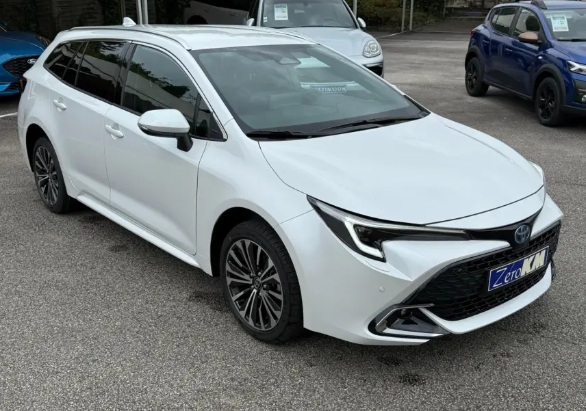 Toyota Corolla Touring Sport blanche nacrée vue en 3/4 avant droit sur parking avec jantes alliage et calandre noire.