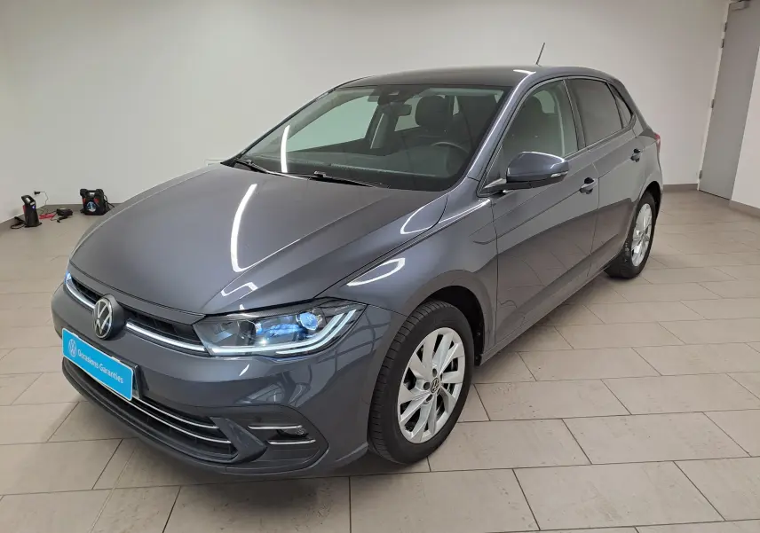 Volkswagen Polo gris cendré en 3/4 avant droit, avec phares allumés et vitres arrière surteintées.